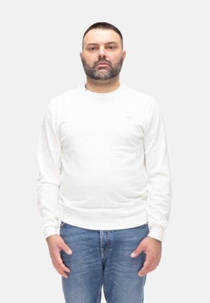 Uomo con capelli corti e scuri e barba, indossa una camicia bianca a maniche lunghe e jeans blu, in piedi davanti a uno sfondo chiaro e semplice.