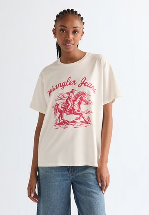 Kobieta z zaplecionymi włosami, ubrana w białą koszulkę Wrangler Jeans z czerwonym nadrukiem jeźdźca na koniu, w połączeniu z niebieskimi dżinsami.