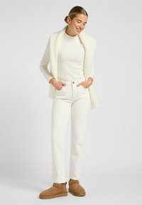 Top de manga larga blanco con costillas, combinado con unos jeans blancos de pierna recta, complementado por un cárdigan oversized de crema texturizada y botas de ante en color beige.