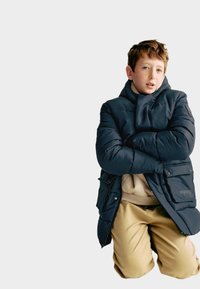 Marineblauwe pufferjack met hoge kraag en twee voorkanten zakken, gedragen over een beige top; kaki broek, met gevouwen armen, neutrale achtergrond.