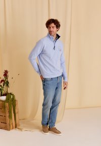 Lichtblauwe pullover met een kwart rits, met een zeilbootafbeelding op de linkerkant van de borst, gecombineerd met blauwe jeans en beige schoenen.