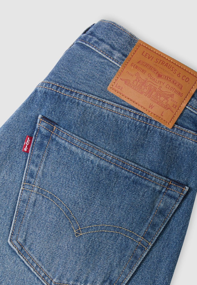 Jeans de mezclilla azul claro con un diseño distintivo en el bolsillo trasero, costuras naranjas y una etiqueta de marca de cuero marrón en la cintura.
