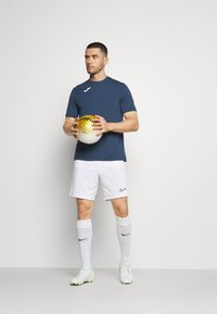 Kék sportpóló logóval, fehér rövidnadrág, fehér zokni és zöld részletekkel ellátott focicipő. Arra tart egy arany- és fehér focilabdát.