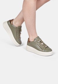 Olive green sneakers with a shiny bronze accent, featuring metallic laces and a white platform sole. Smooth leather texture and streamlined design.