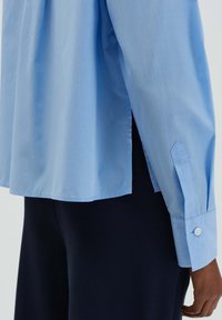 Camicetta azzurro chiaro con una texture liscia, caratterizzata da un taglio dritto, spacchi laterali e un polsino appuntito con bottoni bianchi. Pantaloni scuri parzialmente visibili.