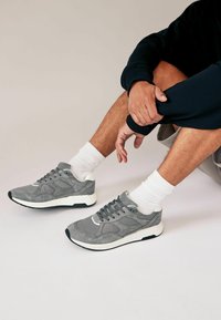 Grå sneakers med en blandning av mocka och mesh, med en vit sula, texturerade detaljer och en klassisk snörningsdesign, kombinerat med vita strumpor.