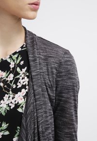Grå strukturerad cardigan med öppen front, lager ovanpå en blommig svart topp som har vita och rosa blommor samt gröna accenter.