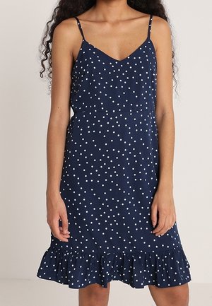 Granatowa sukienka bez rękawów w białe groszki, cienkie ramiączka, dekolt w serek, dopasowana talia i falbanki u dołu, noszona przez stojącą osobę.