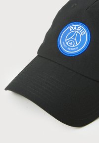 Casquette noire en tissu, ornée d'un patch circulaire bleu avec "PARIS" et "SAINT-GERMAIN" en texte blanc et un élément de design ressemblant à un ballon.