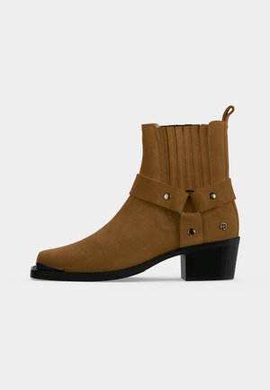 Brauner Velourslederschuh mit mittlerer Schafthöhe, Blockabsatz, elastischen Seitenteilen, Metallnieten und dekorativem Riemen über dem Knöchel.