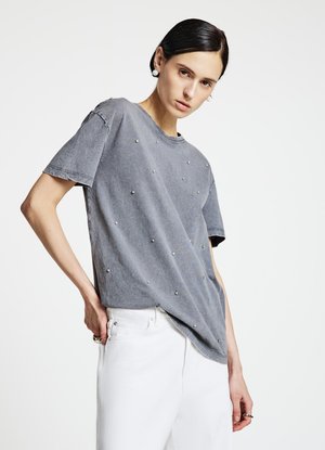 T-shirt grigia a maniche corte con piccoli borchietti decorativi sul davanti. Il tessuto ha una finitura leggermente consumata. Abbinata a pantaloni bianchi.