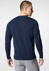 Granatowy sweter z dzianiny z długimi rękawami, prążkowanymi mankietami i dolnym wykończeniem, z okrągłym dekoltem i gładką teksturą. Szare spodnie widoczne poniżej.