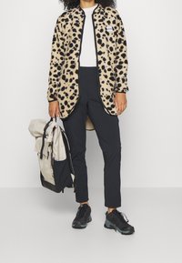 Kabát z fleece s leopardím potiskem a černým lemováním, oblečený přes bílou blůzu a černé kalhoty; nese béžový batoh; sportovní černé tenisky.