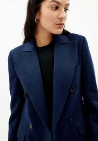 Marinblå dubbelknäppt kappa med stora slag, två framfickor och svarta knappar; tyget verkar slätt och strukturerat.