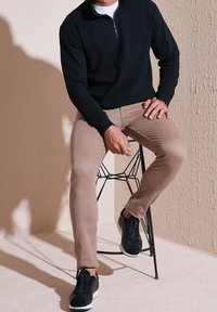 Felpa nera con zip, pantaloni a sigaretta beige e sneaker nere su uno sgabello di metallo contro uno sfondo chiaro e strutturato.
