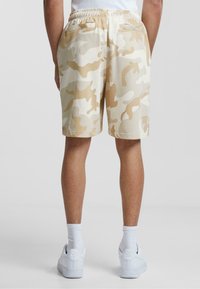 Beige camouflage shorts with an elastic waistband, two back pockets, and a loose fit, paired with white sneakers and socks.