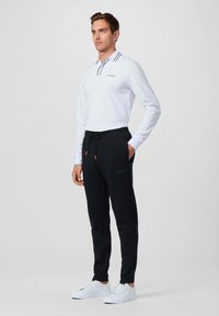 Witte polo met lange mouwen en marineblauwe accenten, gecombineerd met zwarte drawstringbroek met oranje toggles en logo. Model draagt witte sneakers.