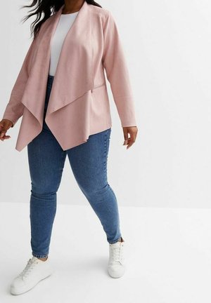 Osoba ubrana w jasnoróżową, otwartą na przodzie drapowaną kurtkę, biały top, niebieskie wąskie dżinsy i białe trampki, stojąca na białym tle.