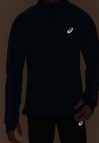 Persona che indossa una giacca e pantaloni sportivi neri con il logo bianco Asics sul petto e sulla coscia.