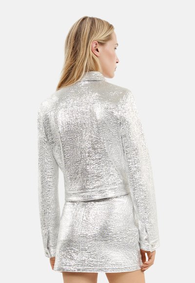 Srebrna metaliczna krótka kurtka o teksturowym wykończeniu i długich rękawach, zestawiona z dopasowaną mini spódnicą. Widok z tyłu pokazuje wyprofilowane szwy.
