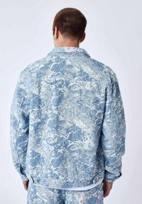 Giacca di denim blu con un motivo astratto e testurizzato, dal taglio leggermente oversize e con chiusure a snap all'orlo. Dettagli di cucitura visibili.