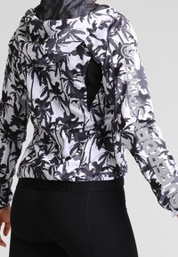 Chaqueta con cremallera en blanco y negro que presenta un patrón de hojas de palma, capucha de malla y el logotipo "SUPERDRY" en la manga. Tejido suave y ligero.