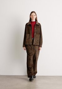 Brown corduroy jacket with two front pockets, worn over a red top and matching brown trousers. Smooth texture with a relaxed fit. Black footwear.