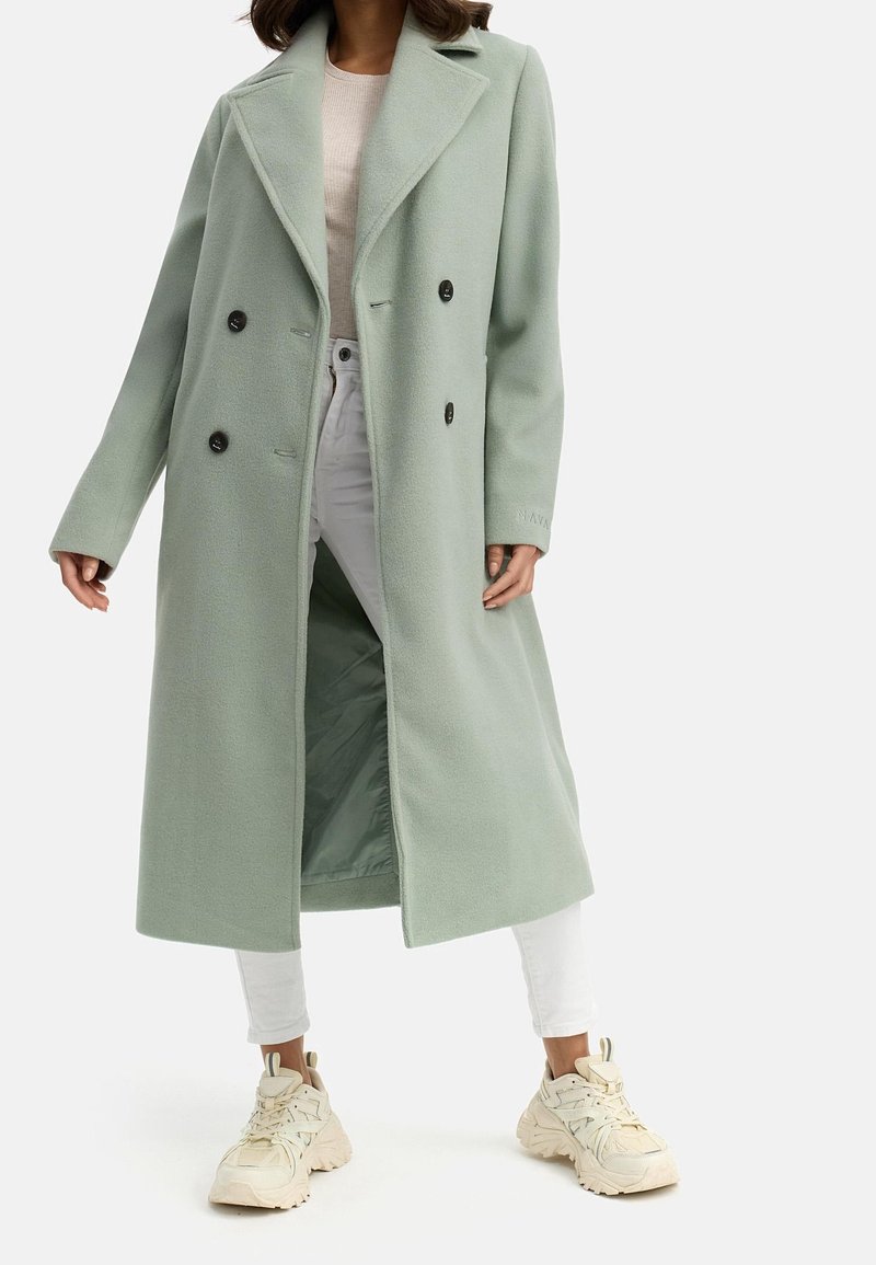 Femme portant un long manteau vert clair à double boutonnage sur un pantalon blanc et des baskets beiges épaisses, debout devant un fond uni.