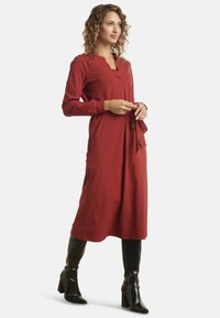 Red long-sleeve dress with a V-shaped neckline, belted waist, and smooth texture, paired with knee-high black heeled boots.