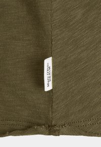 Oliwkowa bawełniana tkanina o prążkowanej fakturze, z białą metką z napisem "Wyprodukowano w Portugalii" oraz tekstem "Denim" przy szwie.