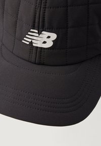 Čierna čiapka vyrobená z prešívanej látky, s zakriveným šiltom a výrazným bielym logom "NB" na prednej strane. Hladký povrch s prešitými detailmi.