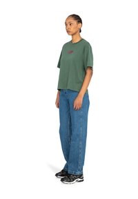 T-shirt verde in cotone a maniche corte con colletto girocollo e logo ricamato, abbinato a jeans blu a vestibilità ampia e scarpe da ginnastica nere.