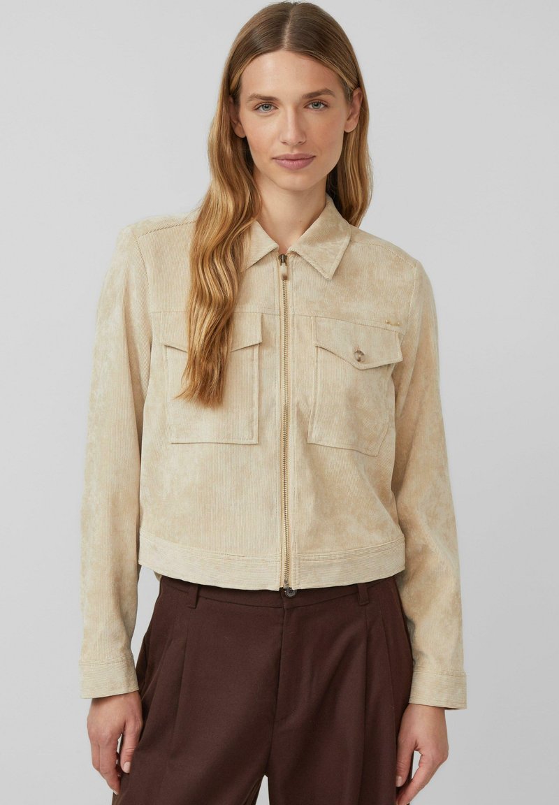 Beżowa kurtka z korduru o kroju krótkim z zapięciem na suwak z przodu, dwoma kieszeniami na piersi i kołnierzykiem, z charakterystyczną fakturą materiału i długimi rękawami.