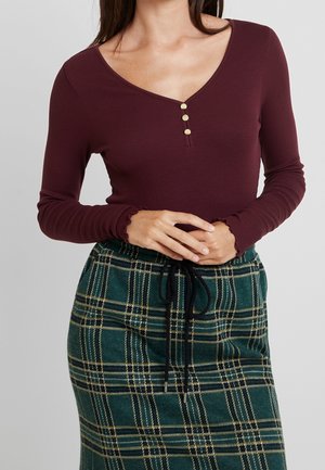 Mujer vestida con una blusa de manga larga en tonos burdeos con una textura acanalada y tres botones, y una falda de cuadros verdes con un cordón negro atado en la cintura.