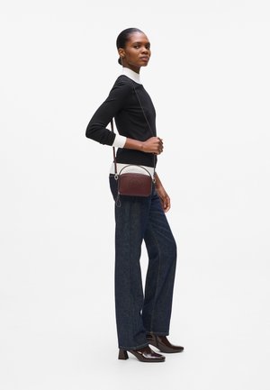 Crossbody bag in burgundy leather with embossed logo, featuring a zip closure and detachable strap. Model wears dark jeans and a black top.