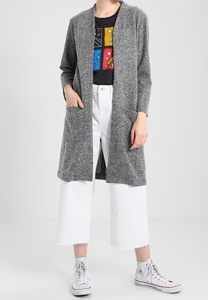 Femme portant un cardigan gris ouvert, un t-shirt noir à motif, un pantalon blanc large et court, et des baskets blanches montantes.