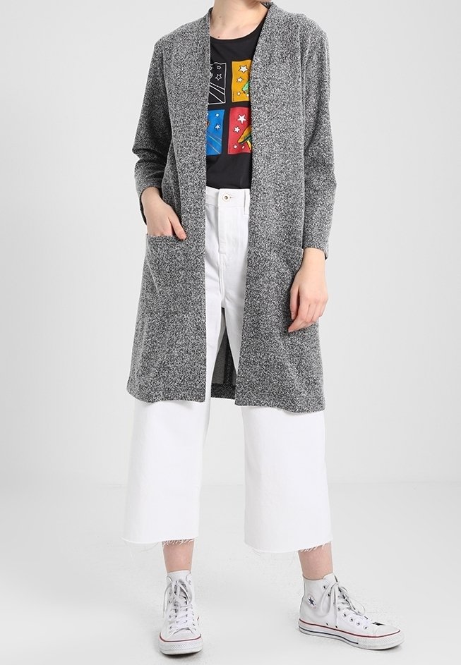 Femme portant un cardigan gris ouvert, un t-shirt noir à motif, un pantalon blanc large et court, et des baskets blanches montantes.