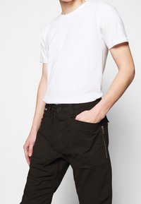 Muž v jednoduchém bílém krátkém tričku s krátkým rukávem, zastrčeném do vysokých černých kalhot s kapsou na zip, s rukou opřenou o přední kapsu.