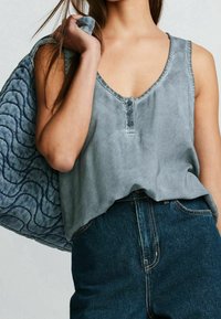 Grå ærmeløs top med V-udskæring og tre knapper foran, sat sammen med mørkeblå højttaljede jeans og en tekstureret skuldertaske.