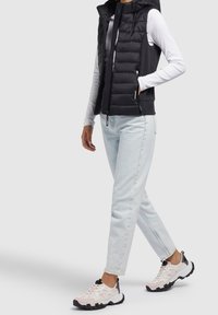 Schwarze Steppweste mit Reißverschluss, kombiniert mit einem weißen Langarmshirt und hellblauen Jeans. Auffällige schwarz-weiße Sneaker.