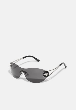 Gafas de sol con lentes gris oscuro y elegante, marco de metal plateado y un acento en forma de estrella negra a cada lado. Diseño contemporáneo curvado.