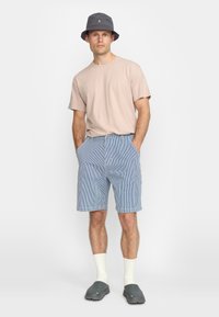Lichtroze t-shirt met korte mouwen, blauw-wit gestreepte shorts, grijze bucket hat, witte kniekousen, grijze klompen, staande pose.
