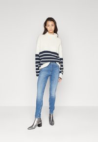 Suéter de punto grueso blanco con rayas marinas, combinado con jeans ajustados azul claro y botines metálicos. Diseño simple y de corte relajado.