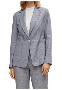 Blazer grigio con texture, a collarino sceso, chiusura a un bottone e due tasche anteriori. Abbinato a un top leggero in maglia.