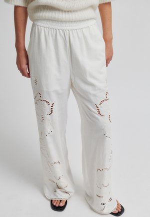 White wide-leg pants with floral cutout embroidery worn with a white sweater and black open-toe sandals on a plain background.