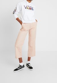 Sudadera blanca con el logotipo "VANS" en impresión multicolor. Pantalones de pierna ancha en rosa claro. Zapatillas negras con acentos blancos y un calcetín. Diseño simple.