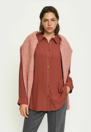 Vrouw draagt een roestkleurig overhemd met knopen, een roze pluizige sjaal over haar schouders, grijze broek, staand tegen een effen achtergrond.
