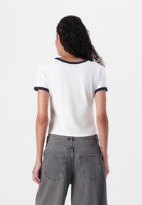 White cotton t-shirt with navy blue ribbed trim at the neckline and sleeves, paired with high-waisted gray denim pants featuring back pockets.