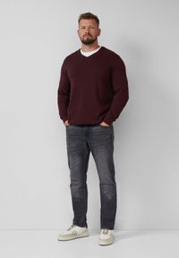 Burgunderfarbener V-Ausschnitt-Pullover, kombiniert mit dunklen Jeans mit verwaschenen Details und hellen Sneakers in Creme. Das Model steht mit den Händen in den Taschen.