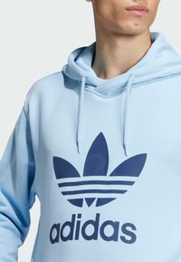 Felpa con cappuccio di colore azzurro chiaro, caratterizzata da un grande logo trefoil di adidas in navy e strisce orizzontali sul fondo. Tessuto morbido.